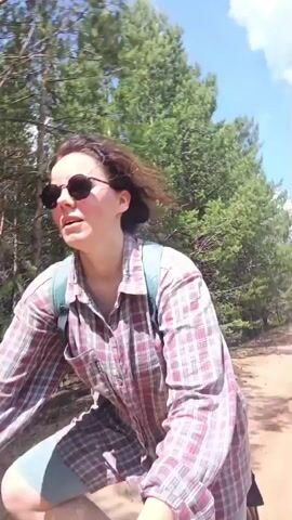 A young woman with curly brown hair and sunglasses, wearing a pink and white plaid shirt and a teal backpack, rides a bicycle along a dirt path. She is smiling and looking to her left, with her mouth slightly open as if speaking. The path is lined with tall green pine trees and the sky is blue with a few white clouds. The camera is positioned low and to the side, giving a dynamic perspective of the cyclist and her surroundings.