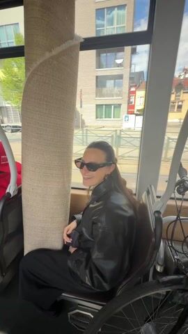 A woman sits in a transit vehicle, possibly a tram or bus. She is wearing sunglasses and a black leather jacket with a black top and black pants. Her hair is pulled back with a gold-colored hair clip. She is seated near a large roll of carpet. She looks out the window and then down at the carpet. In the background, other passengers are visible, as well as buildings through the windows. The transit vehicle appears to be moving, based on the changing scenery visible outside.