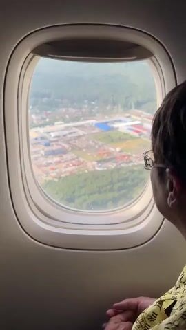 This is a POV video from an airplane window. A person wearing glasses and a yellow shirt is looking out the window at a suburban landscape with buildings and trees below. The plane appears to be in the process of ascending or descending, as the ground is visible through the window. The footage is slightly shaky, indicating movement of the camera.
