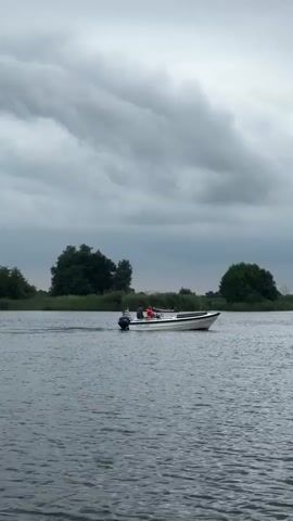 A white boat with two people on board travels across a wide, calm body of water. The sky above is overcast with dark, cloudy conditions. The boat is moving from left to right in the frame. The water is a deep gray color, reflecting the cloudy sky. On the far bank, there is a line of green trees and tall reeds. The boat appears to be a small motorboat. The two individuals in the boat are wearing life vests. One person is seated at the helm. The boat is leaving a slight wake behind it.