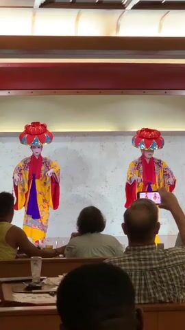 Two performers in traditional Japanese attire with vibrant yellow kimonos and large red headdresses are on a stage. The performer on the left has their arms outstretched and hands open, while the performer on the right is holding a small item and looking down. Several audience members are seated at tables in the foreground, with one person in the center foreground filming the performance with a smartphone. The stage has a textured white backdrop and is lit from above. The overall environment appears to be an indoor dining area.