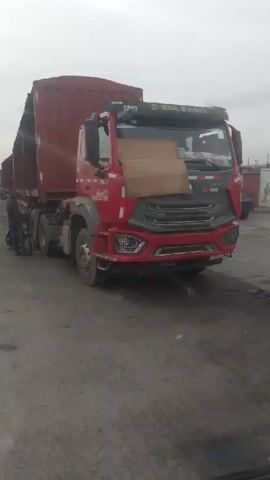 A person is leaning down to work on the front passenger side wheel of a red semi-trailer truck. Two other people are standing nearby, one of whom is also leaning down near the wheel. The truck is parked on a paved surface with other trucks lined up in the background, suggesting a truck yard or a roadside rest stop. The sky is overcast. The truck has a red cab with a tan piece of cardboard taped to the front windshield. The truck is hauling a large red container.