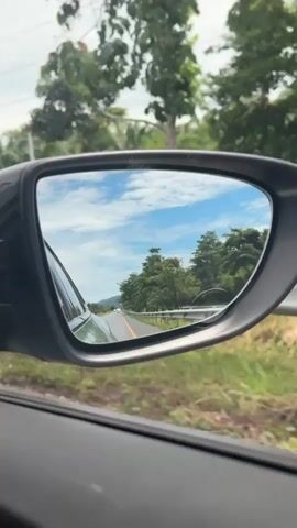 The video shows a side-view mirror of a car driving on a road surrounded by lush greenery and trees under a cloudy sky. The reflection in the mirror displays the road ahead, with other vehicles visible in the distance. The car's interior is visible on the left side of the frame. The scene suggests a journey through a tropical or sub-tropical landscape.