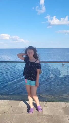 A young girl stands on a paved walkway next to a body of water. She is wearing a dark, off-the-shoulder shirt with white polka dots and light blue shorts. Her hair is long and dark, and it blows in the wind. She is wearing purple shoes. The water is a deep blue, and there are white clouds in the sky. A glass railing separates the walkway from the water. The girl is looking off to the side.