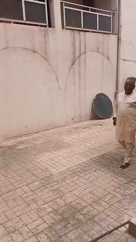 This media file captures a moment in Sabon Gari North, Nigeria. The scene is set outdoors, paved with brickwork, adjacent to a light-colored building. A man in traditional attire, a light-colored kaftan and cap, is walking. He appears to be smiling. He is accompanied by two individuals in camouflage uniforms, suggesting they are security personnel. The person in the foreground of the uniform is wearing a hat and is looking towards the man in traditional attire. The sky appears overcast, suggesting it might be daytime with cloudy weather. The building has windows with railings and a balcony. The overall atmosphere seems formal, possibly indicating an official movement or escort. No visible text is present in the media.