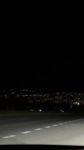 The video depicts a night scene from a roadside perspective. The foreground shows a road with white dashed lines, illuminated by headlights. In the distance, a city is visible with numerous lights scattered across the landscape.