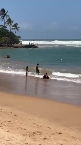 The video shows a beach scene on a sunny day. The foreground is a sandy beach. In the mid-ground, the ocean meets the shore with gentle waves. Several people are visible in the water and on the beach. In the shallow water near the shore, two children are playing. One child is sitting on the sand in the water, and another child, a boy, is standing nearby, facing the other child. A third child, a girl, is standing to the left of the boy, also facing the child in the water. Further out in the ocean, more people are swimming and enjoying the water. In the background, there is a rocky outcrop covered in vegetation and palm trees. More people are visible on the rocks and in the water near the rocks. The ocean stretches to the horizon where large waves are breaking. The sky is blue with a few scattered clouds. The overall impression is one of a lively and pleasant beach environment.