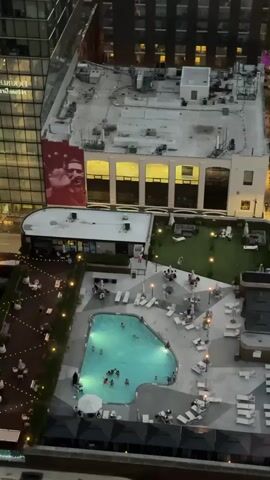 This is an aerial shot of a rooftop pool area. The pool is illuminated with turquoise lights, and several people are swimming in it. Around the pool are lounge chairs and umbrellas. To the left of the pool is an area with tables and chairs, possibly an outdoor dining space. Beyond that, string lights are visible. To the right of the pool is a patch of artificial turf with more lounge furniture. In the background, a large building has a mural of a man on its side. The scene appears to be a lively social gathering space.