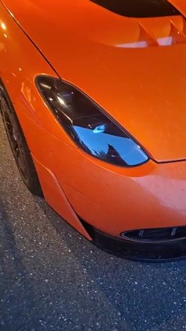 A close-up shot shows the front passenger side of an orange sports car. The car has black accents on the hood and a black front bumper. The headlight is visible, reflecting a blue light. The camera pans slightly to the right, revealing more of the car's body and a black barrier in the foreground. The background is dark, with metal trusses and lighting fixtures overhead.