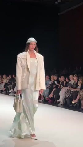 A fashion show is taking place on a runway with an audience seated on either side. A female model, wearing a white blazer over a white dress with a subtle pattern, walks down the runway. She is also wearing a white headscarf and carrying a white handbag. The audience consists of men and women, some of whom are looking at the model, while others are using their phones. The background is dark, with some lighting from the runway and stage.