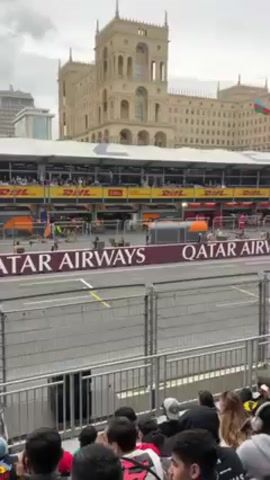 The video shows a Formula 1 race car speeding past a crowd of spectators. The car is green and silver, and it is moving very fast. The spectators are sitting in the stands, and they are cheering as the car passes. Behind the racetrack is a grandstand with “DHL” and “Qatar Airways” written on the facing. Behind that is a large tan building with multiple towers. There is an Azerbaijan flag blowing in the wind. The weather is overcast.