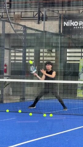 A young man wearing a black sleeveless shirt and black pants is playing padel. He is in the process of hitting a yellow padel ball with a black padel racket. Several other yellow padel balls are visible on the blue court, lined up near the net. The court is enclosed by glass walls and a net. In the background, a sign reads "Niko Padel Academy." The environment appears to be an indoor sports facility.