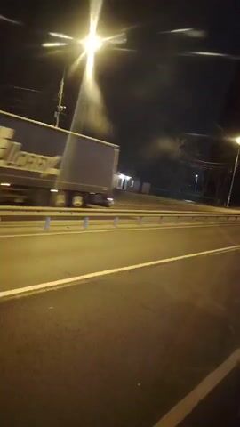 A night time video shows a road with a guardrail. A lamppost shines brightly overhead. A white trailer truck passes by on the road.