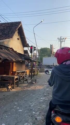 A motorcycle rider wearing a pink helmet and a gray hoodie is stopped at a red traffic light on a dirt road. To the left of the road is a small, rustic building with a corrugated metal roof, appearing to be a shop or food stall. Several wooden tables and benches are set up outside the building. Beyond the traffic light, the road continues, with other motorcycles and a car visible. The environment appears to be a rural or semi-urban area with overhead power lines and trees in the background. The lighting suggests it is either early morning or late afternoon.