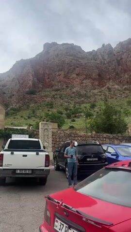The video shows an exterior view of a stone church and a large, rocky mountain in the background under a cloudy sky. In the foreground, there are several cars parked on a paved lot, including a white pickup truck, a black SUV, a red sedan, and a white sedan. People are walking around the cars and near the entrance to the church grounds. Some individuals are seen entering or exiting through a stone archway. A man in a blue shirt is standing near the black SUV, holding a cup. Another man and a child are near the white sedan.