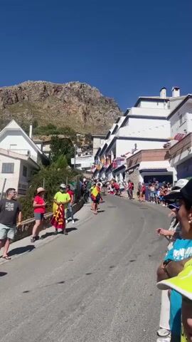 The video shows a street lined with white buildings with blue trim, decorated with flags of various countries. A steep, paved road curves upwards into the distance. On the sides of the road, a crowd of people are gathered, many wearing casual clothing. Some individuals are wearing or holding flags, including Spanish flags. A banner with Spanish text is visible hanging across the street. The scene appears to be a festive gathering or event, possibly a race or procession, given the upward sloping road and the spectators. The sky is clear and blue, indicating a sunny day. A rugged, rocky mountain forms the backdrop to the buildings. People are standing, walking, and observing the scene. There is a sense of anticipation or excitement among the crowd.