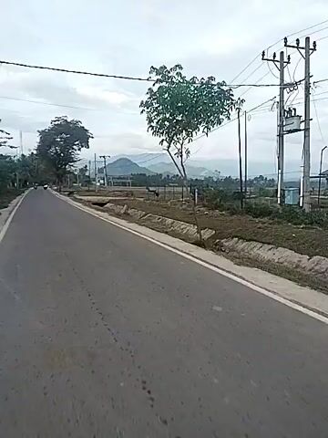 The video shows a street. A person on a scooter passes the camera. Mountains are in the distance. The sky is overcast. Trees are alongside the road. There is tall grass and drainage ditches near the road. Power lines run alongside the road.