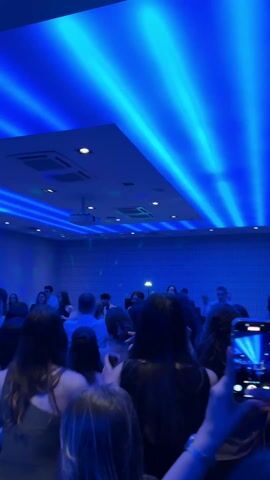 A low-angle shot displays a crowd of people, mostly facing away from the camera, in what appears to be a dance or party setting. The room is illuminated with vibrant blue lighting, which creates linear patterns on the ceiling and walls, giving the impression of a modern or club-like atmosphere. A projector can be seen attached to the ceiling. Several people are actively dancing with their hands up, while one individual is holding up a smartphone, filming the event. The camera captures the scene as a concert video, with the phone's screen mirroring the blue lights and the crowd. The focus remains on the crowd and the light display.