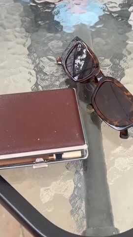 A hand with beige nails and a black ring opens a brown leather cigarette case on a glass table. Inside the case are rows of brown and white cigarettes held in place with a black elastic band. The hand closes the case. A pair of brown sunglasses sits to the right of the case. The table is outdoors.