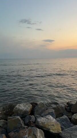 The video captures a serene ocean view at dusk. The foreground is dominated by a cluster of large rocks, appearing to be part of a shoreline or breakwater. The ocean stretches out in front, its surface rippling gently. In the distance, the sky displays the soft hues of sunset, with pastel colors blending into a pale blue overhead. A few wispy clouds dot the horizon. At the 00:04 mark, a small wave crashes against the rocks, creating a brief splash. No people or animals are visible in the frame.