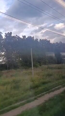 The video shows a view from a moving vehicle, likely a train, looking out a window. The scene outside is rural, with a dirt path or track in the foreground. A grassy field with scattered yellow flowers stretches into the middle ground, bordered by a dense line of trees. In the background, a large metal telecommunications tower, with a lattice structure and antennas at the top, is visible. The sky is partly cloudy, with sunlight breaking through. Power lines are strung across the top of the frame. The perspective shifts slightly as the vehicle moves, suggesting a continuous journey. There are no people visible in the frame.