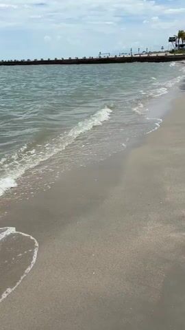 The video shows a view from a sandy beach towards the ocean. Small waves gently lap onto the shore. In the distance, a long pier or seawall extends across the horizon, with structures visible on it. The sky is partly cloudy. The water is a greenish-blue color, and the sand is light brown. The camera is positioned low, near the water's edge, looking out towards the sea and the distant pier. There are no people or animals visible in the frame.