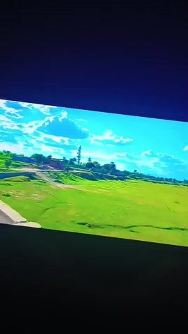 A video shows a bright green grassy field with trees and buildings in the distance, under a blue sky with white clouds. The video is displayed on a rectangular screen in a dark environment.