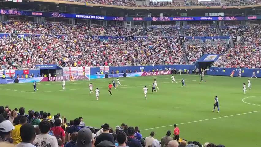 A wide shot shows a soccer match being played in a packed stadium. The field is green, with white lines marking the boundaries. Players in white and blue uniforms are visible on the field. The stands are filled with a diverse crowd of spectators, many of whom are wearing casual clothing. The upper sections of the stadium display banners advertising "FIFA WORLD CUP 2026" and "MIAMI." The lower sections of the stadium have advertisements for "QATAR AIRWAYS" and "WORLD'S BEST AIRLINE." The camera is positioned in the stands, behind a group of spectators, providing a view of the game from a distance. The sky is overcast.