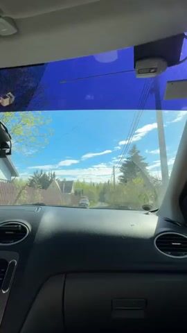 The video shows a drive down a narrow, slightly overgrown residential street. The view is from inside a car, looking through the windshield, which has a blue tint at the top. The sky is blue with scattered clouds.  There are houses on either side of the street, surrounded by trees and bushes.  Utility poles and wires run alongside the road. A car is visible in the distance. The dashboard of the car is black and shows air vents and a radio. The sound of music can be heard.