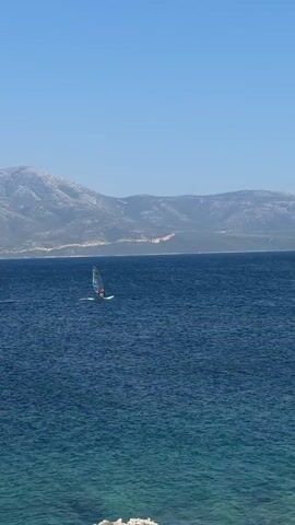 A lone windsurfer is shown on a calm, blue ocean. The windsurfer is wearing a red top and a white life jacket. They are positioned on a white board with a sail that is predominantly white and blue. The windsurfer is in the middle of the frame, relatively small against the vastness of the water. In the background, a mountain range with visible slopes and some scattered vegetation stretches across the horizon under a clear, bright blue sky. The water in the foreground has a slightly lighter turquoise hue, suggesting shallow water or a different lighting condition, with a hint of white foam from small waves. The overall scene depicts a peaceful day at sea with a single individual engaged in a water sport.