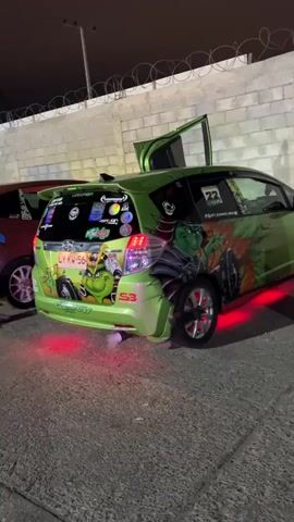 A lime green Honda Fit is parked at night, its rear doors and the driver's side door are open. The car is heavily customized with various stickers on the rear windshield and body, including a large graphic of a cartoon turtle character on the rear quarter panel. The car has custom red underglow lighting. The license plate reads LY KW 56. The car appears to be at an outdoor venue, with a concrete wall and chain-link fence in the background. A dark cable is visible on the ground to the right of the vehicle. The car is emitting a puff of white smoke from its exhaust pipe.