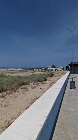 This is a vertical video clip showing a beach scene in Ömirzaq, Kazakhstan. The time of day appears to be late morning or early afternoon, as indicated by the bright sunlight and clear blue sky with scattered clouds. The weather is sunny and warm, typical of a summer day.

The foreground of the video is dominated by a sandy beach with sparse, dry vegetation. A white concrete barrier runs along the right side of the frame, separating the sandy area from a paved walkway. Beyond the beach, the calm, blue sea stretches to the horizon.

In the middle ground, there are several rows of beach umbrellas, mostly green and yellow, providing shade for beachgoers. People can be seen walking along the shore and near the umbrellas, suggesting a relaxed atmosphere and a day for leisure. Further in the distance, there are some structures that might be beach facilities or small buildings.

In the far background, the water meets the sky, and on the right side of the frame, a modern street lamp is visible, as is a trash bin. The overall impression is one of a pleasant day at the beach, with a mix of natural scenery and recreational amenities. There is no visible text in the video.