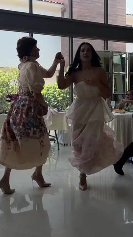 Two women are dancing indoors in front of a large window. One woman is wearing a strapless, layered white dress with pink floral accents. The other woman is wearing a light-colored dress with puffy sleeves and a floral skirt. They are both wearing high heels. The scene appears to be a celebration or a party, with tables set up in the background. The light coming from the window suggests it is daytime. The women seem to be enjoying themselves, with smiles and energetic movements. The floor is reflective, indicating a polished surface.