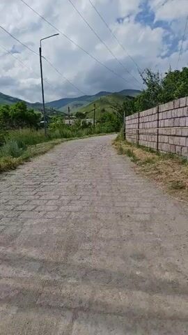 This is a vertical, first-person perspective video clip of a paved road in Bazum, Armenia. The road is made of grey cobblestones and is flanked by dry grass and a stone wall on the right. In the distance, rolling green hills rise under a cloudy sky. Several utility poles with wires stretch across the frame, and a modern street lamp stands on the left side of the road. The overall atmosphere is calm and serene, suggesting a peaceful rural setting. The time of day appears to be daytime, with the sun occasionally breaking through the clouds. No visible text is present in the clip. The scene depicts a person walking down the road, presumably on their way to a cafe to get coffee.