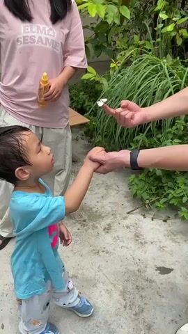 A toddler in a blue shirt and pajama pants reaches out his left hand. A person with an arm wearing a black smartwatch gently places three small white butterflies onto the toddler's open palm. Another person wearing a pink t-shirt with "ELMO SESAME" visible on it and light-colored pants stands in the background holding a yellow bottle. The ground is a concrete or stone surface, and there is lush green foliage behind the people.