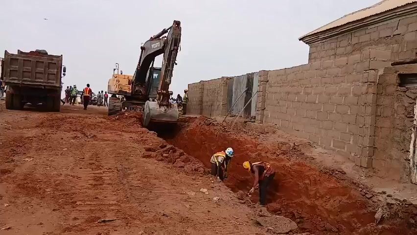 The scene depicts a construction site where an excavator is digging a trench in reddish soil. Two workers wearing hard hats and safety vests are working within the trench with shovels. A dump truck loaded with soil sits nearby, and a crowd of people watches from the edge of the site. A brick wall borders the construction area. The sky is overcast.