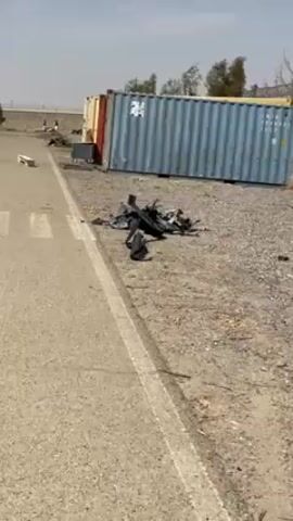 The video begins with a close-up of debris, mostly black and silver, lying on dirt and gravel. A shipping container stands in the background. The camera pans to show a road with trees lining the side. Buildings with white roofs are visible in the distance beyond a fence, and a few people are standing in that area.