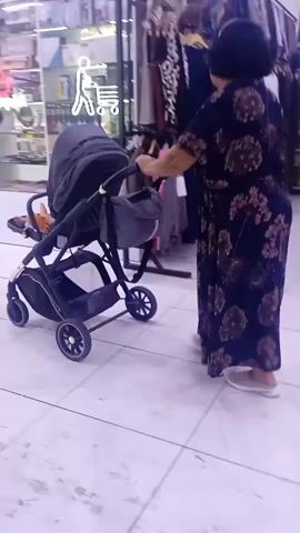 In a brightly lit, indoor marketplace in Ak-Suu, Kyrgyzstan, a woman in a dark floral dress pushes a stroller. The floor is tiled with a checkerboard pattern of white and light gray tiles, and overhead fluorescent lights illuminate the space. To the left, shelves are stocked with various goods, some of which are obscured by displays. To the right, clothing racks are visible, displaying a colorful assortment of garments. The woman is the central focus, her back to the camera as she navigates the aisle with the stroller, which has a black bassinet and a dark gray storage bag.

Further down the aisle, another woman and a young boy are walking towards the camera, engaged in conversation. The woman is wearing a black t-shirt and blue jeans, while the boy is in a blue t-shirt and jeans. Behind them, other shoppers can be seen walking through the marketplace. The atmosphere appears to be that of a typical busy shopping day, with no indication of specific weather or time of day beyond the artificial lighting. The overall scene conveys a sense of everyday commerce within a local market setting.