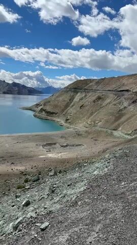 The video pans across a large, tranquil lake with vibrant turquoise water. Majestic, rugged mountains rise in the background, partially shrouded by dramatic clouds under a bright blue sky. The foreground is characterized by a barren, rocky shoreline and a dusty, dry landscape. The overall scene depicts a remote and breathtaking natural environment.