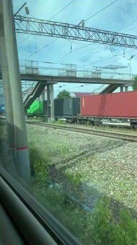 A train car window reveals a view of a train carrying cargo containers on parallel tracks. The containers are of various colors, including green, red, black, and blue. Overhead, power lines and a pedestrian overpass are visible. The ground beside the tracks is covered in gravel and some green vegetation. The video appears to be recorded from another moving train.