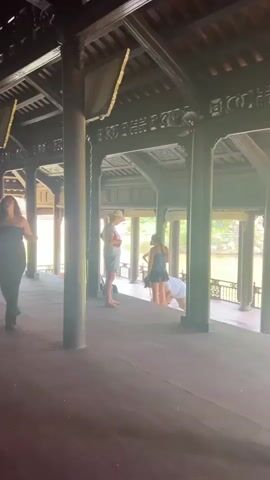 The video shows a wooden structure with columns and a roof. Sunlight streams through the open sides, illuminating the interior and casting shadows. Three people are visible. One person, wearing a hat and light-colored clothing, stands near the left side of the frame, looking towards the right. To their right, another person in dark clothing is bending down, seemingly looking at something on the floor. Further to the right, a third person is kneeling down, with their back to the camera. Beyond the structure, a body of water with a yellowish-green tint is visible, bordered by railings and greenery. The overall scene suggests an outdoor historical or cultural site.
