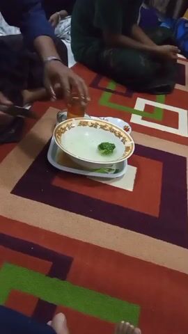 The video shows a low-angle shot of a group of people sitting on a colorful patterned carpet. In the center of the frame, a white tray holds a bowl of light-colored liquid with a single green leaf floating in it, and a small glass of amber liquid. To the left of the tray, a person's arm and hand are visible, wearing a bracelet. To the right, two people are seated, one wearing a green garment. The carpet has geometric patterns in shades of red, brown, green, and white. The overall lighting is dim. The scene appears to be indoors.