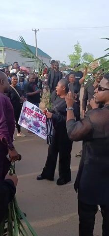 Here is a description of the video:

A group of people are gathered outdoors on a paved surface that appears to be a street. They are holding signs, some of which read "A CRY FOR HELP STOP BENUE KILLINGS" in bold letters. The signs also depict what seems to be bloody scenes. The individuals are dressed in casual attire, with many wearing black clothing. Some people are holding palm fronds, while others are recording the scene with their mobile phones.

In the center of the group, a woman in black is actively speaking or leading the group. Other people are listening or reacting to her. The background shows buildings, utility poles, and what may be a marketplace. The weather seems to be overcast.
