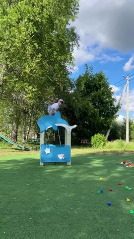 The video shows a young child jumping off playground equipment. A child wearing a white shirt, shorts, and a pink hat climbs on top of a blue and white play structure shaped like a dolphin. The playground is covered in green artificial turf, with scattered colorful blocks nearby. Trees surround the playground on a partly cloudy day. The child stands on top of the play structure and jumps off, landing on the ground. The child kneels on the ground after landing. A slide is visible in the background to the left and utility poles are visible in the background to the right.