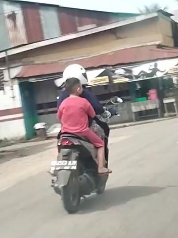 A motorcycle is being ridden by two individuals on a paved road. The rider, in a dark blue top and patterned pants, wears a white helmet. A child, in a red t-shirt and red shorts, sits in front of the rider, also wearing a white helmet. The child is barefoot. The motorcycle has a license plate that reads "B 3503 AJF". Behind the motorcycle, there is a roadside building with a metal roof and a sign that says "25.000". There are trees and vegetation visible in the background. The time stamp indicates the video is from 00:00 to 00:05.