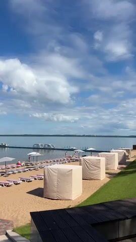 The video shows a sandy beach with numerous lounge chairs and umbrellas set up. In the background, a calm body of water with a pier and a few boats is visible under a partly cloudy blue sky. Several white cabana-like structures are positioned on the sand closer to the camera. A few people are scattered on the beach, some sitting on the lounge chairs, and one person appears to be in the water near the pier. The scene is peaceful and suggests a resort or recreational area.