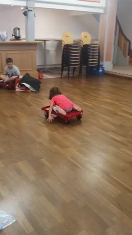 Two children are playing in a room with a wooden floor. One child, wearing a pink shirt and striped pants, is lying on a red flat cart with wheels, pushing themselves forward. Another child, wearing a grey shirt, is sitting on a similar red cart behind the first child. There is a counter in the background with some items on it, and stacks of chairs are visible. The scene appears to be a recreational activity.