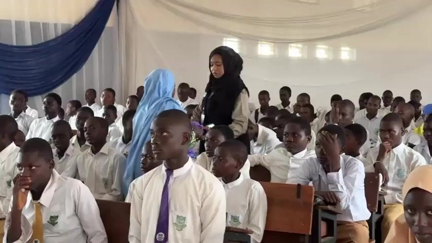 The video depicts an indoor gathering of numerous students, predominantly young males, dressed in school uniforms consisting of white shirts with ties (some purple, others green/yellow). Female students are also present, wearing yellow tops, blue skirts, and hijabs in light brown or light blue, or abaya-like garments. Most students are seated at wooden desks, showing attentiveness as they listen to an unseen speaker whose voice discusses "discipline" and "the ability to discipline yourself."

Two adult females stand towards the rear of the room; one wears a black hijab, the other a light blue one. The setting appears to be a school hall or auditorium in Unguwan Dosa, Nigeria, decorated with white ceiling drapes and a prominent blue fabric draped across the back wall. Papers are visible on some desks, and the students' white shirts feature a green and white crest logo. The atmosphere is focused and engaged, suggesting an educational event or lecture.