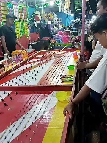 The video shows people playing a carnival game involving a game board with small pegs and balls, which are being dropped onto the board in an attempt to get them into colored bowls. A vendor wearing a black t-shirt and cap stands behind the game, overseeing the proceedings. Prizes such as snacks, small bottles of liquid, and stuffed animals are displayed on shelves behind the game. The environment appears to be a busy carnival or fairground.