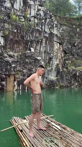 A young man stands on a bamboo raft in a body of green water. He is shirtless and wearing camouflage shorts. He has tattoos on his left arm. Behind him is a large rock cliff face with visible striations. The man performs various dance moves and poses for the camera.